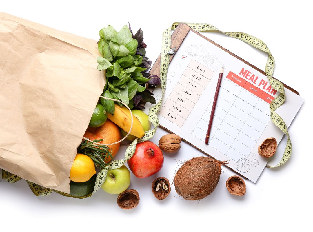There is a brown paper bag with health foods spilling out. On the other side is a clip board and pen with a paper that has a meal plan sheet on it.