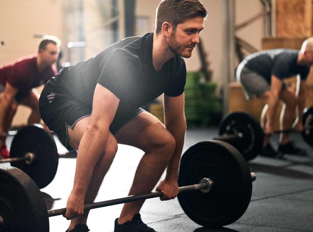 There is a man about to lift up a bar with weights on both sides. He is wearing black and grey clothes and there are two individuals behind him also about to lift the weight bar. They are in a gym.