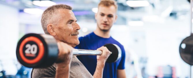 A man with grey hair is lifting a weight bar and a guy in a blue shirt is watching from behind.
