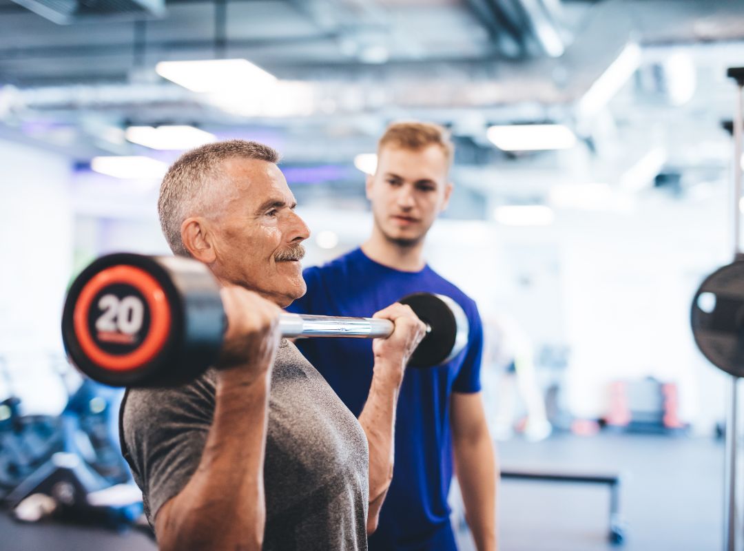 A man with grey hair is lifting a weight bar and a guy in a blue shirt is watching from behind.