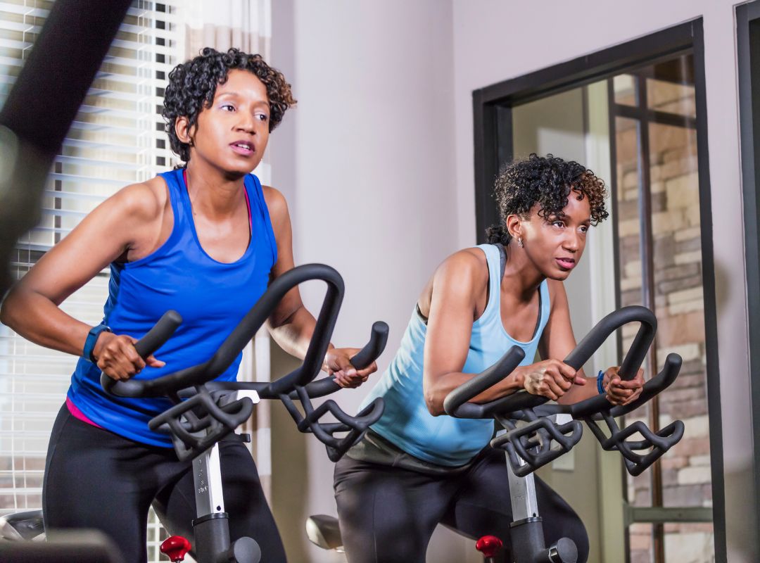 There are two African American women on stationary bikes with blue tops. They have determined expressions on their faces and have light grey walls behind them.