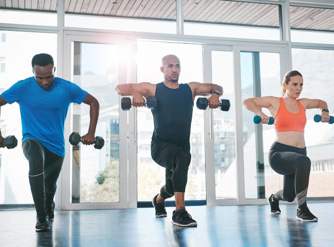 There is a group of people lifting weights. A man on the left is wearing a blue shirt and black pants, the middle man is wearing all black clothes, and the right woman is wearing an orange sports bra and black leggings. They are inside with glass doors behind them.