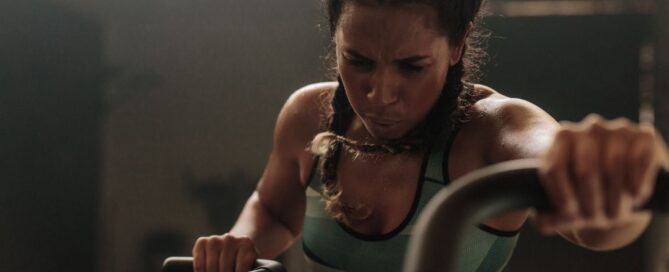 A woman has two braids in her hair and she is wearing a green sports bra and leggings. She is in a dark room and is on an elliptical machine.
