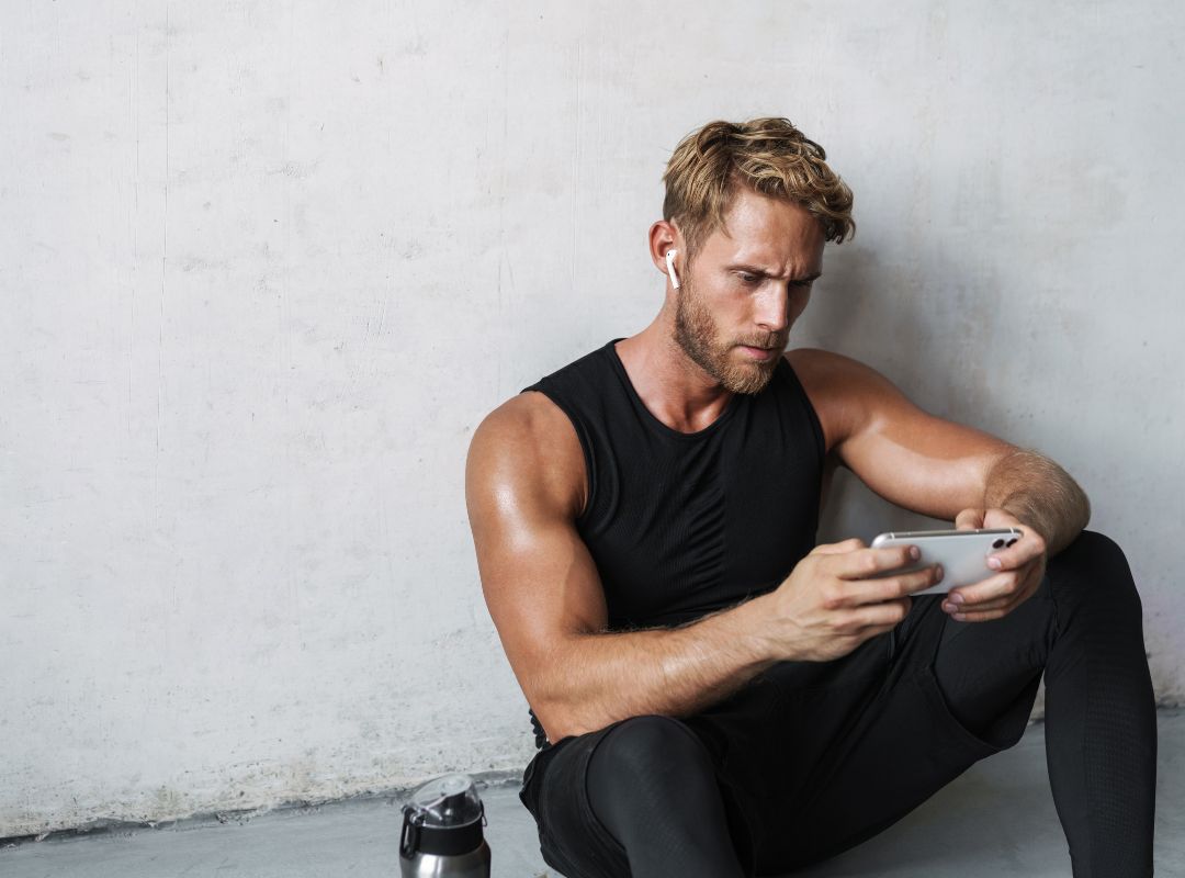 There is a guy in black workout clothes sitting on the floor next to a white wall. He has a metal water bottle next to him and is holding a white phone and has white earbuds in. He has blond hair and is looking at the phone.