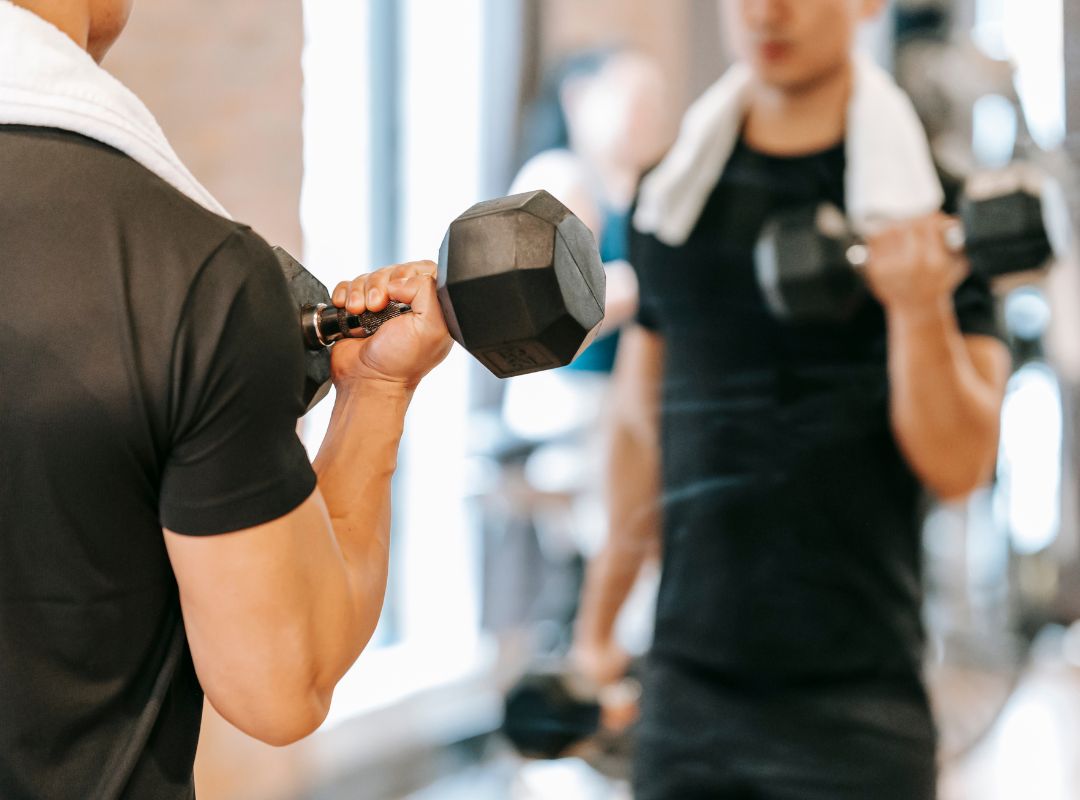 There is a man lifting a weight wearing all black with a mirror in front of him. A woman is also working out in the background out of focus and the man has a white towel on his neck.