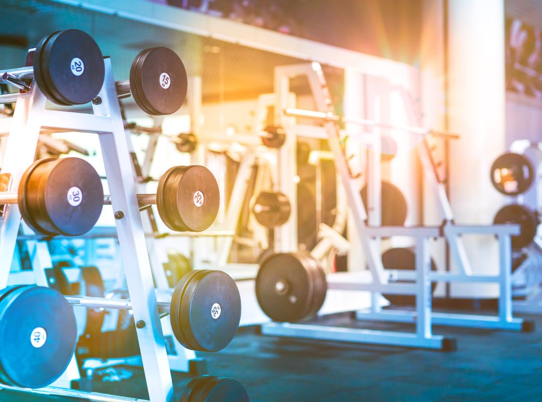 A gym is shown and racks filled with weights are close to the image and there is a mirror on the wall. There is also a light flare over the whole image with orange and blue colors.